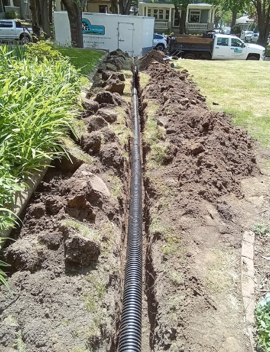 A corrugated black drainage pipe sits inside a narrow, freshly dug trench in a residential yard.