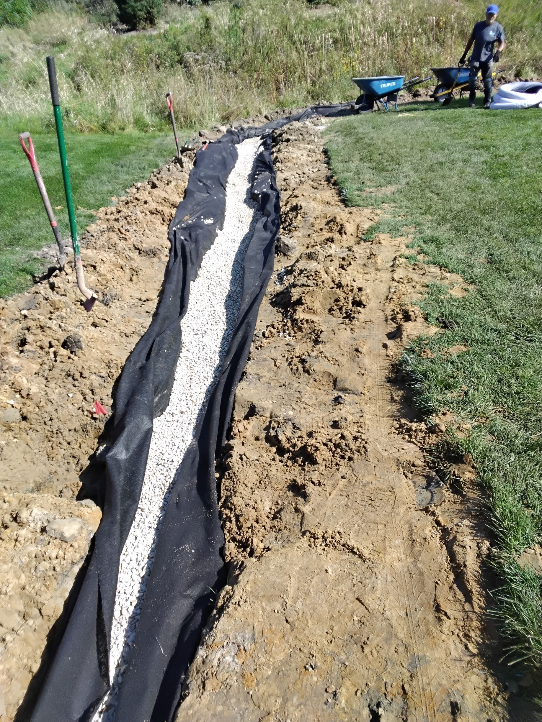 Crew backfilling a French drain pipe with drainage rock