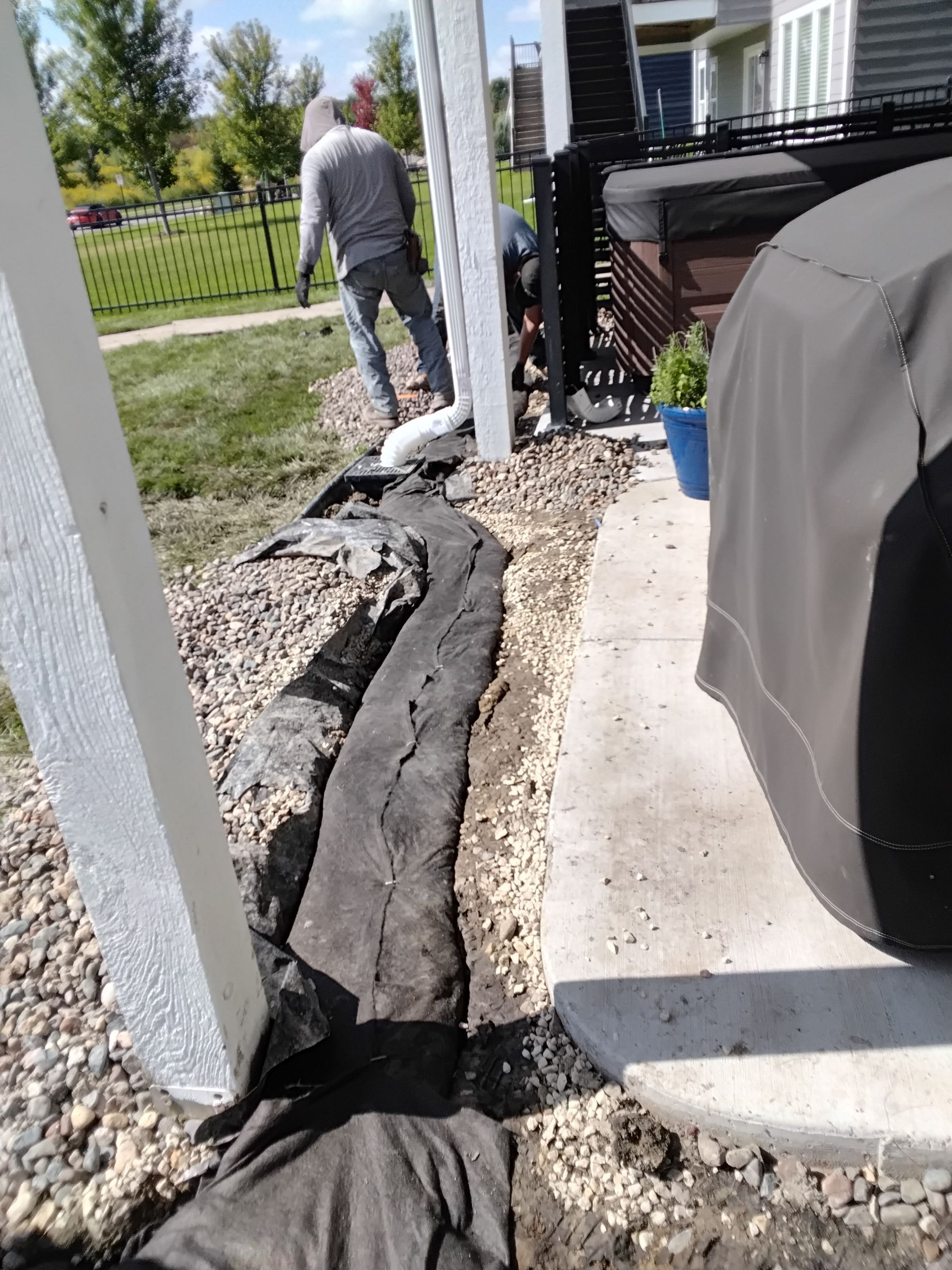 A person in a gray shirt works on a landscape drainage project with black fabric and gravel next to a patio and house.