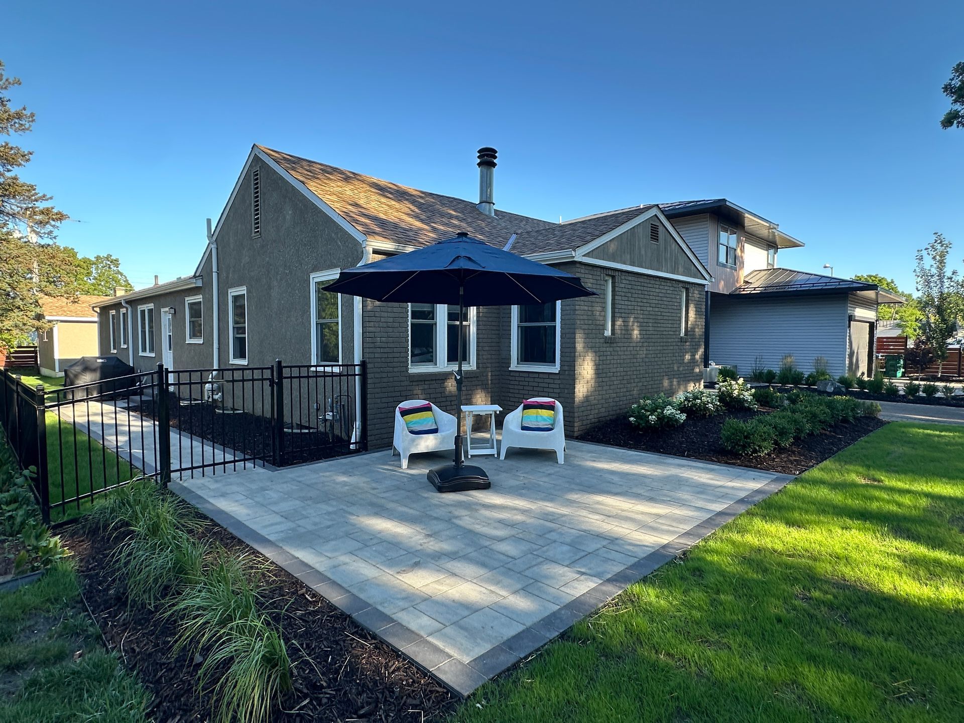 A stone house features a patio with two white chairs under a dark blue umbrella, next to a black fence and green lawn.