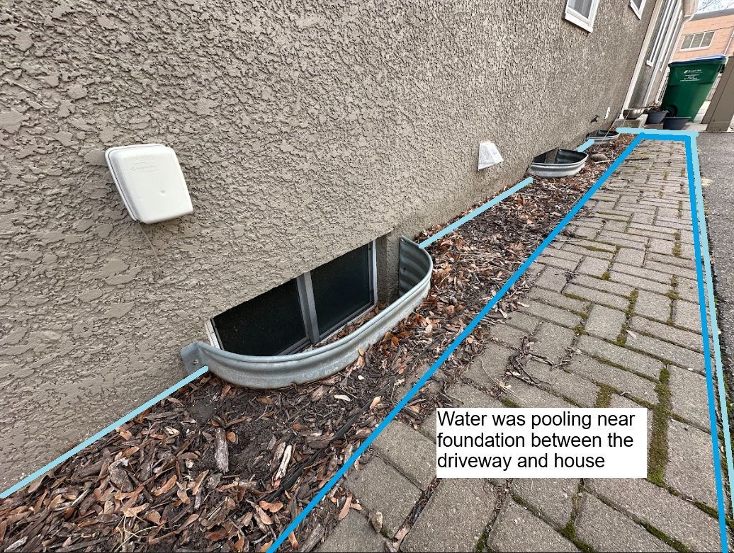A house foundation with a window well, bordered by a paved walkway where a blue line marks an area of water pooling.