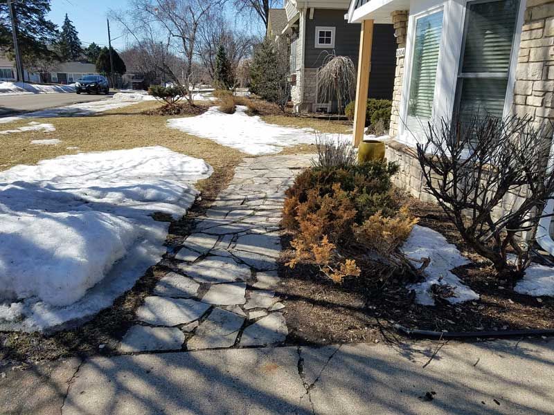 Stone walkway with melting snow on either side, leading to a house with a bay window.