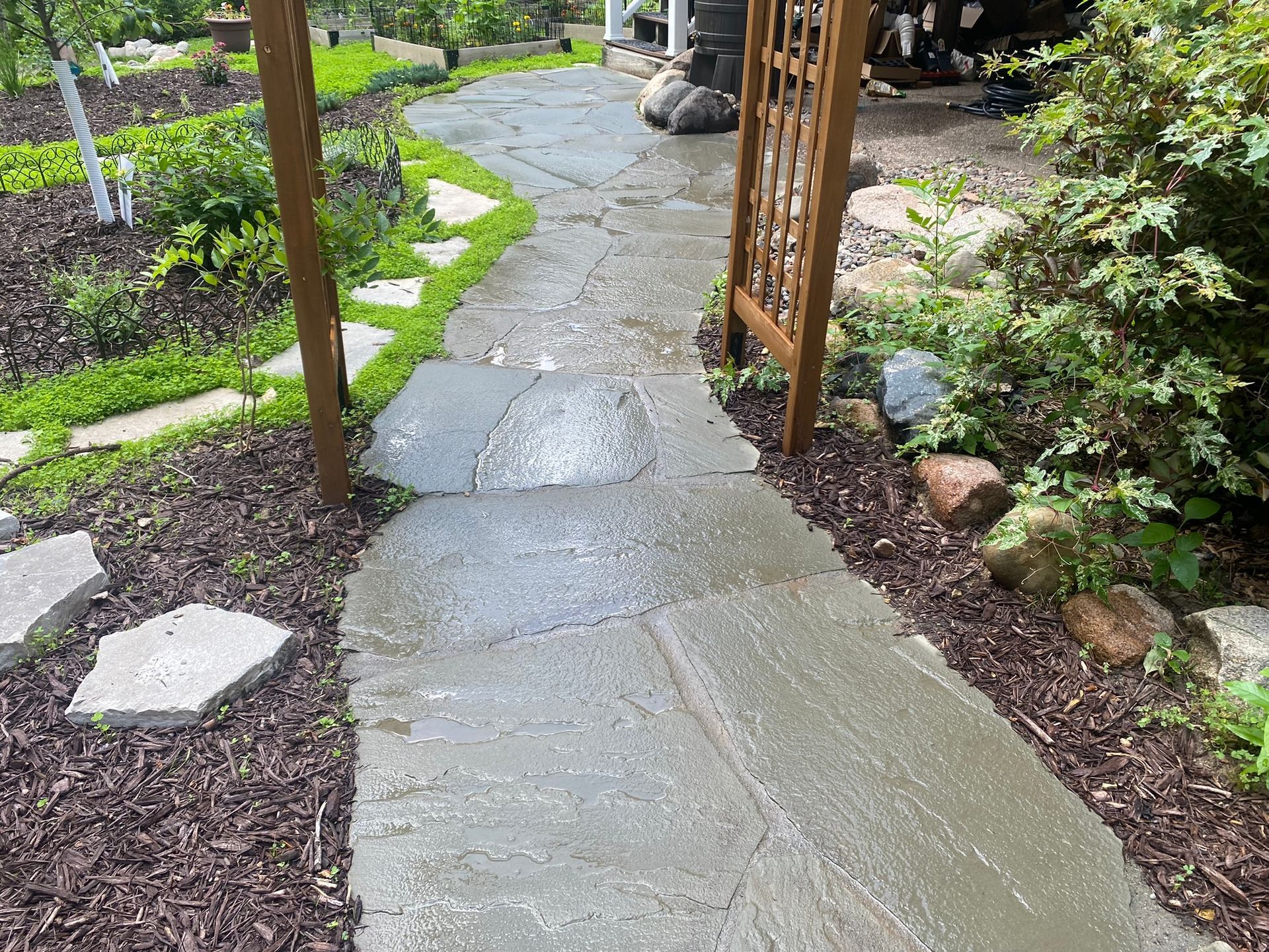 Stone pathway through a garden, leading to a wooden gate. The path is wet and surrounded by mulch and greenery.