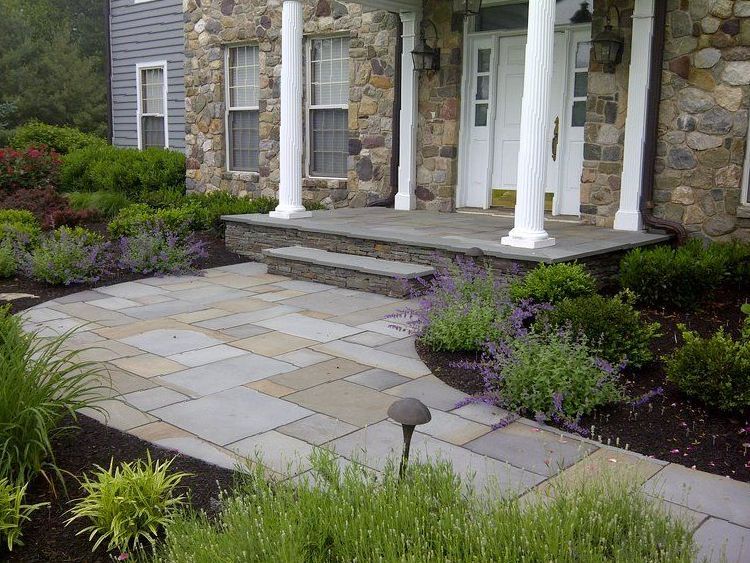 A stone walkway leading to a house surrounded by trees and bushes.