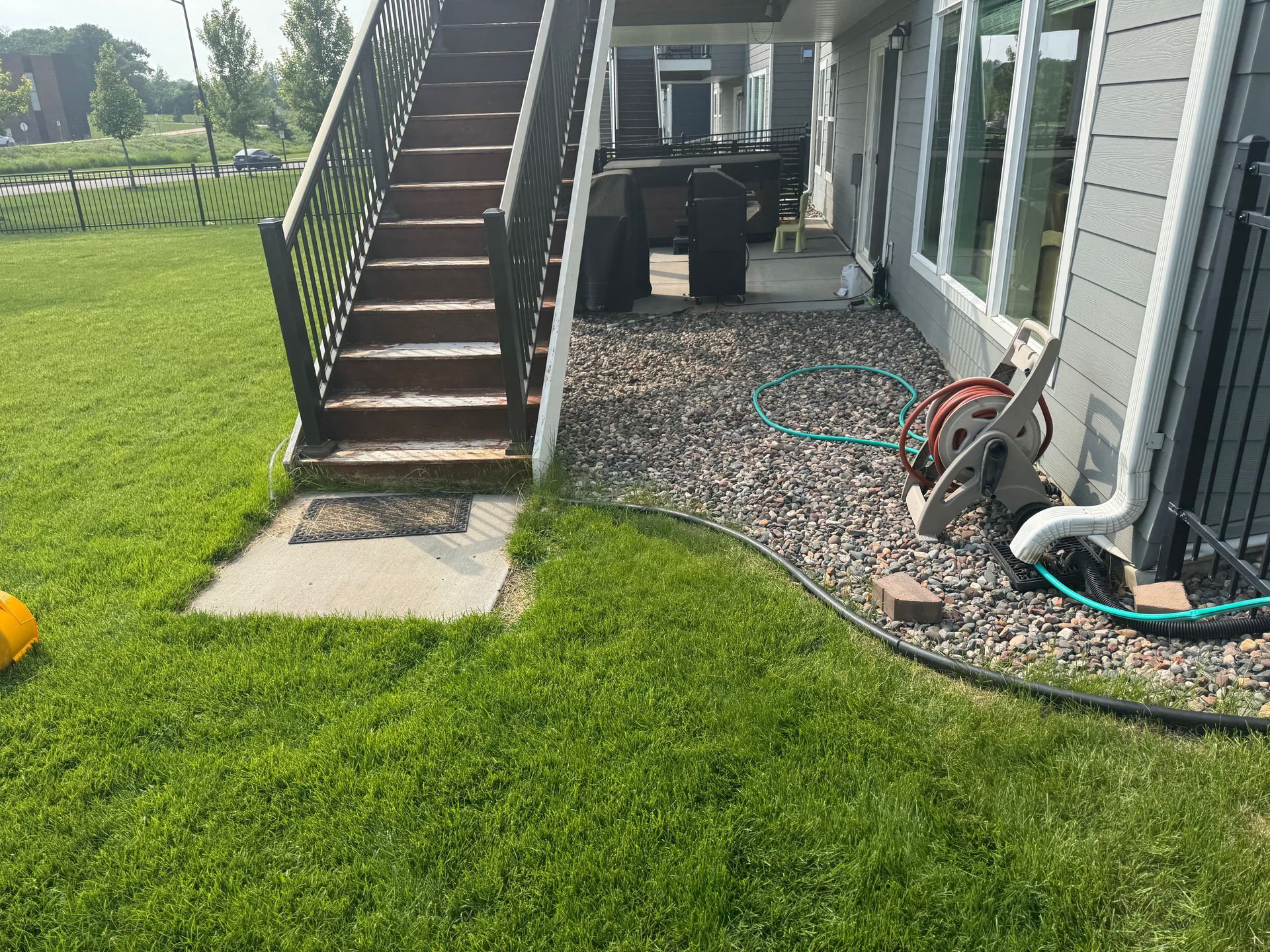 A set of outdoor stairs leads to a covered patio with a gravel ground, green lawn, and a garden hose reel on the side.