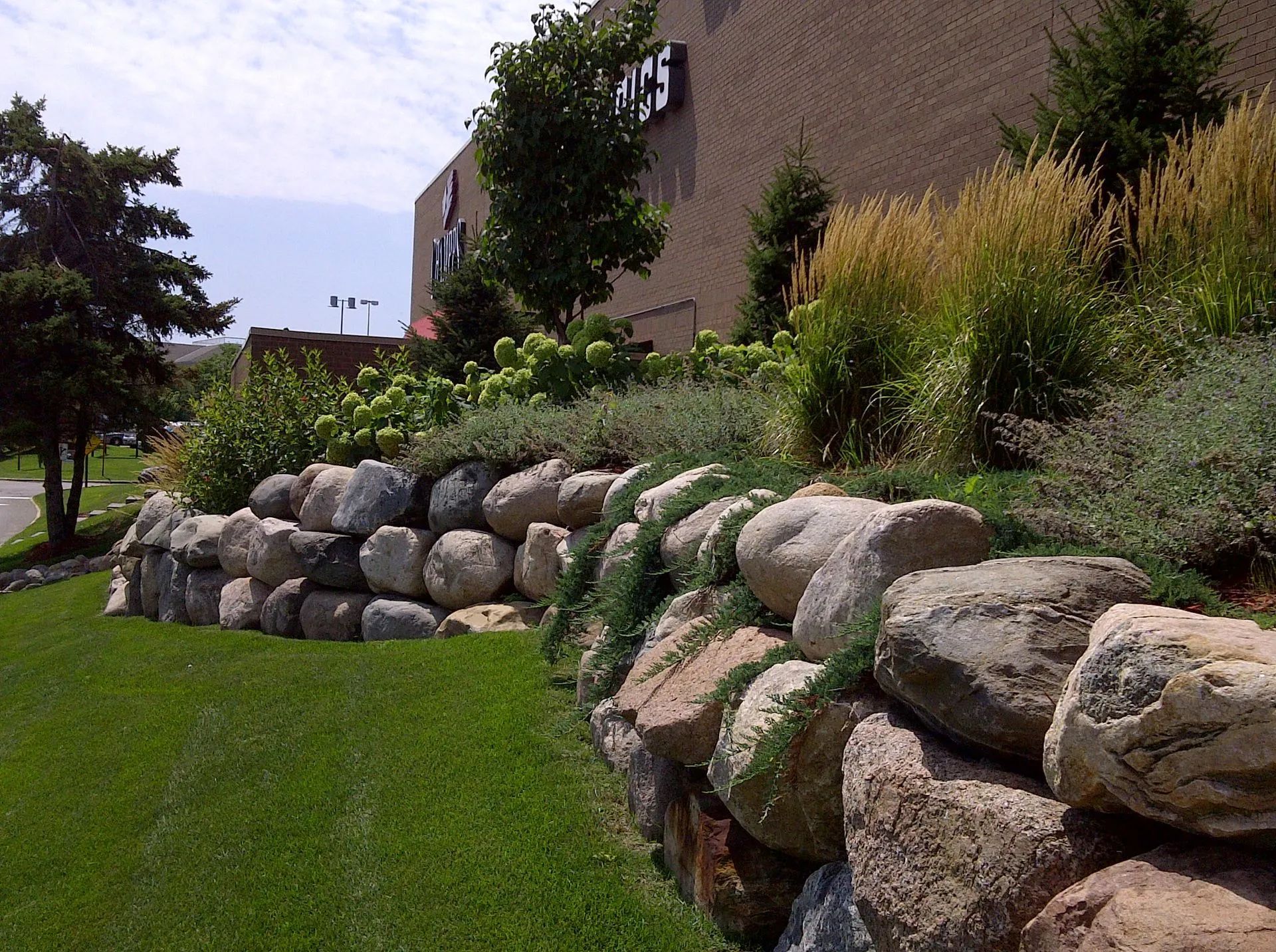 A stone retaining wall in front of a building with grass and greenery on a sunny day.