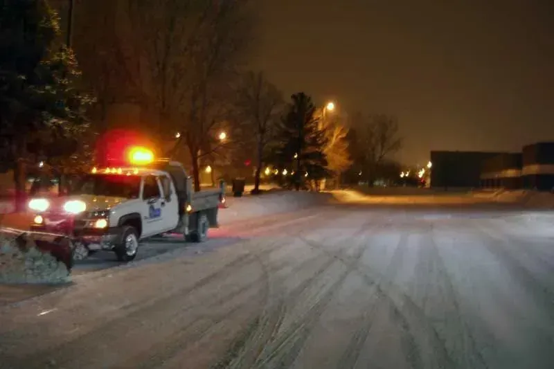 Snowplow clearing a snow-covered street at night, illuminated by streetlights and the truck's flashing lights.