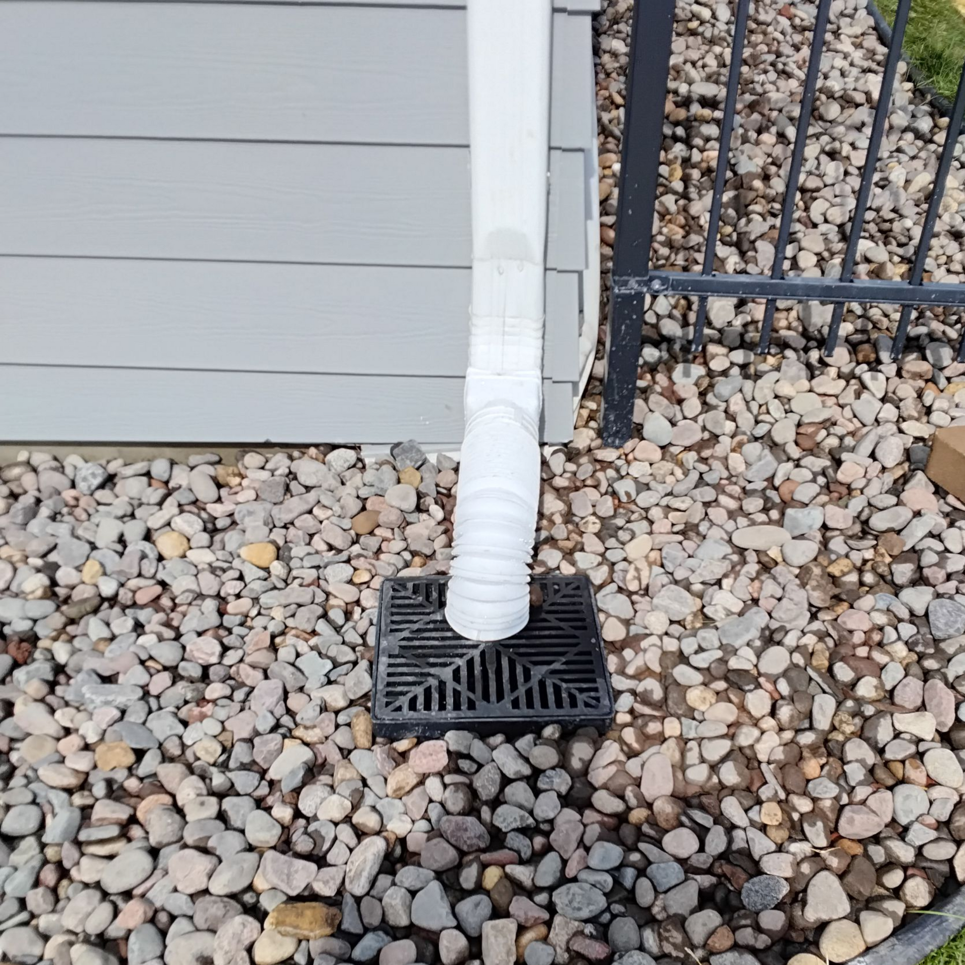 A white gutter downspout extends from the side of a grey house and flows into a black square drain set in gravel.