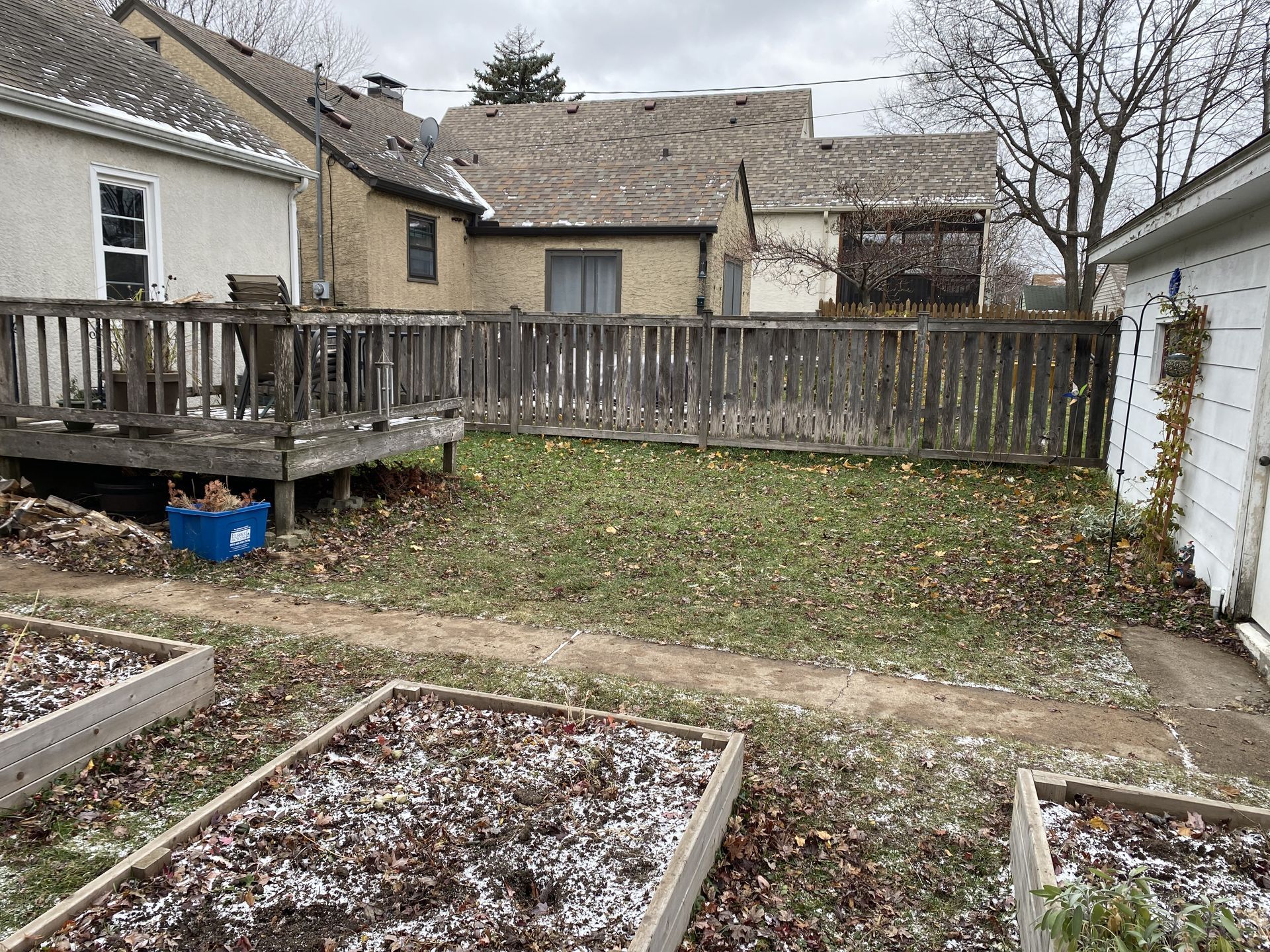 A backyard with a wooden deck, a wood fence, and several raised garden beds covered in light snow.