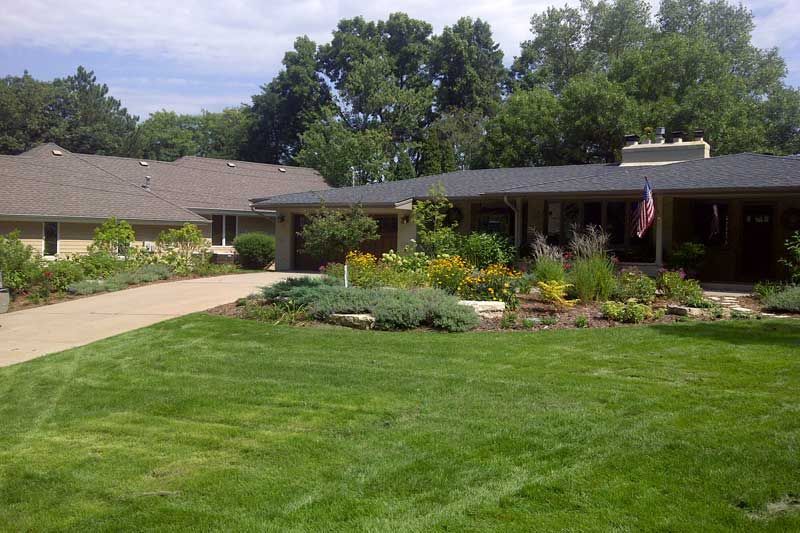 A well-manicured lawn leads to a house with a garden, trees in the background, and an American flag.