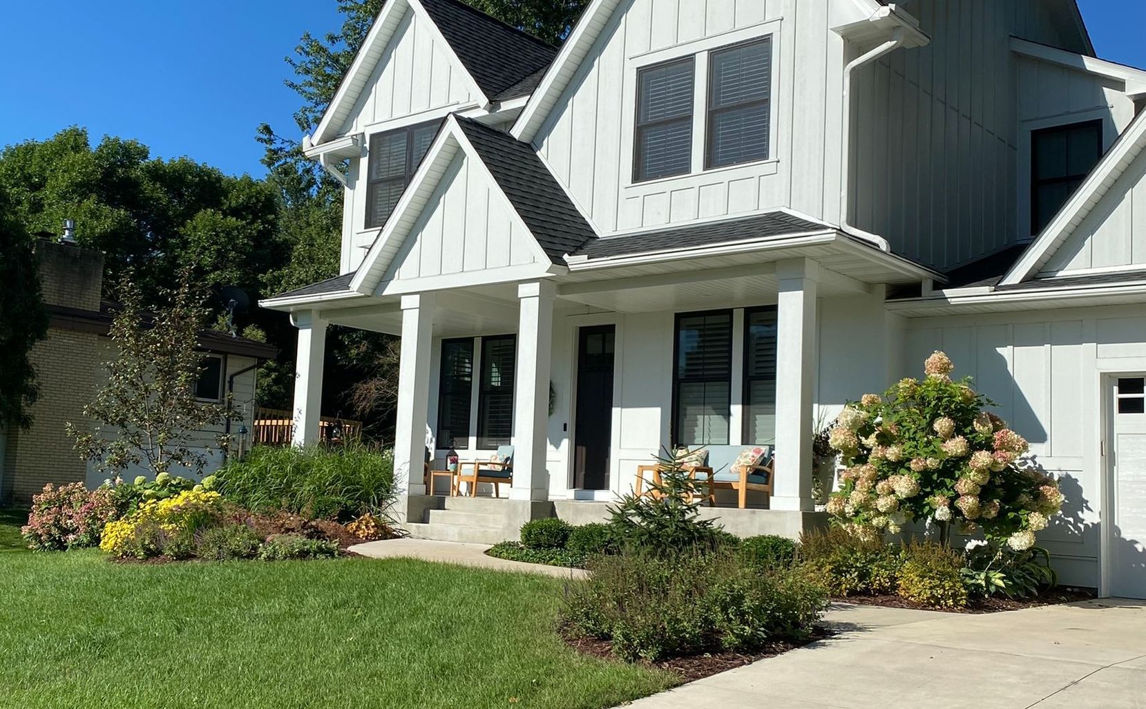 White farmhouse with black shutters, green lawn, and flowering bushes.