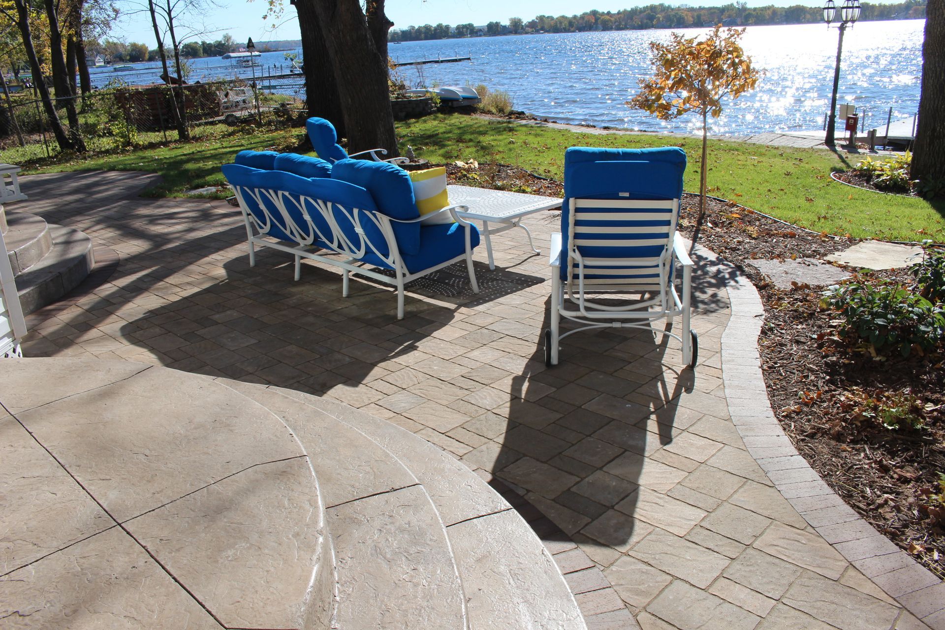 A patio with white metal outdoor furniture and blue cushions, overlooking a lake with trees and green grass.