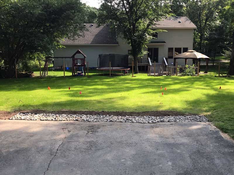 Backyard with a lawn, playground, trampoline, and gazebo, behind a house, with a stone border in front.