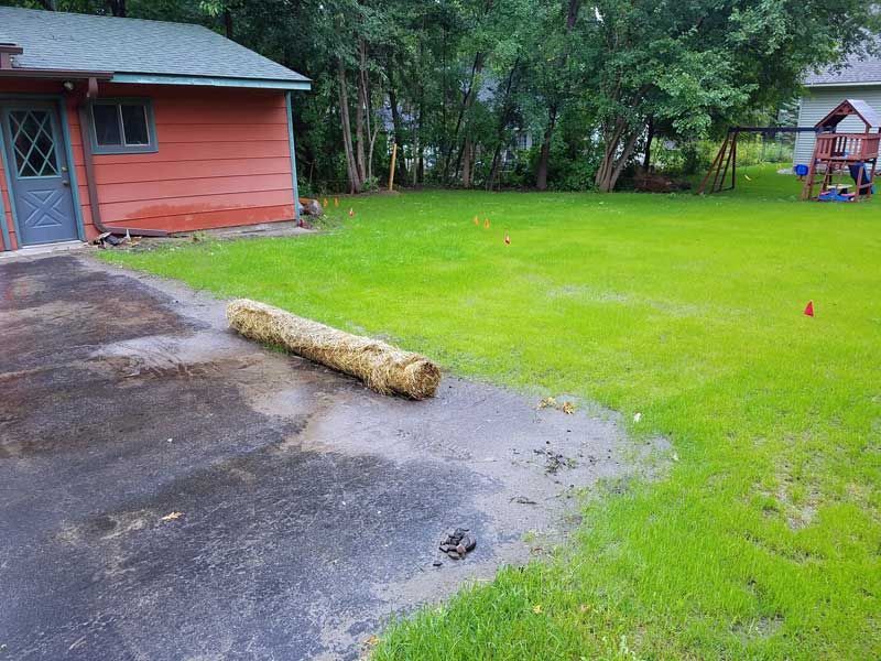 A tan straw log on wet ground next to a red building and green lawn.