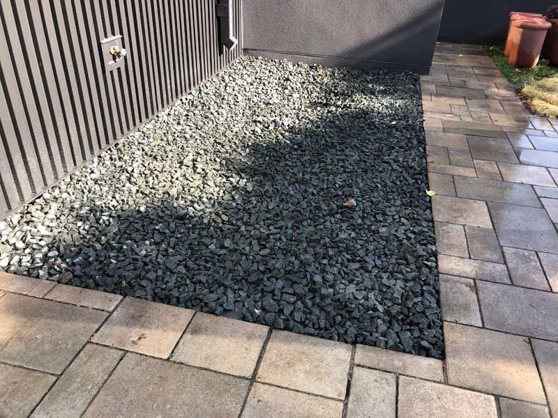 Gravel bed next to a building, bordered by pavers. The ground is covered in dark gray stones.