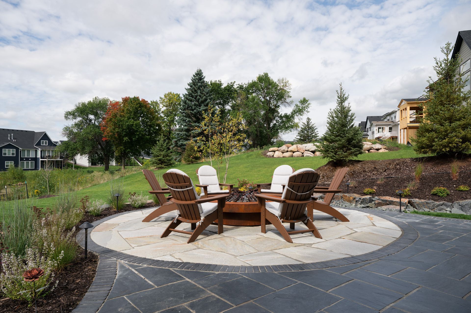 Raised circular flagstone patio with gas fire feature and Adirondack chairs with landscaping filled with boulders and flowers