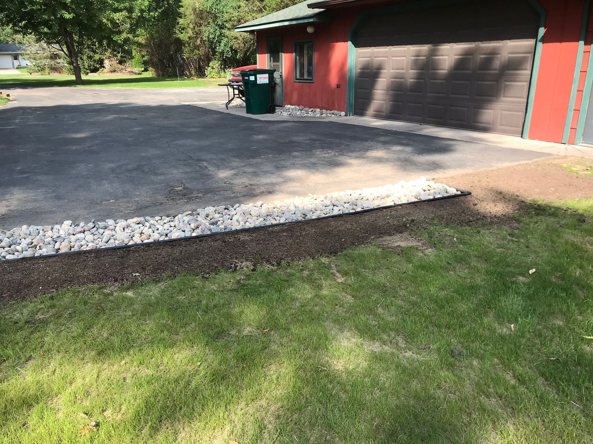 Asphalt driveway next to a red building with a garage. White rocks line the edge, beside grass.