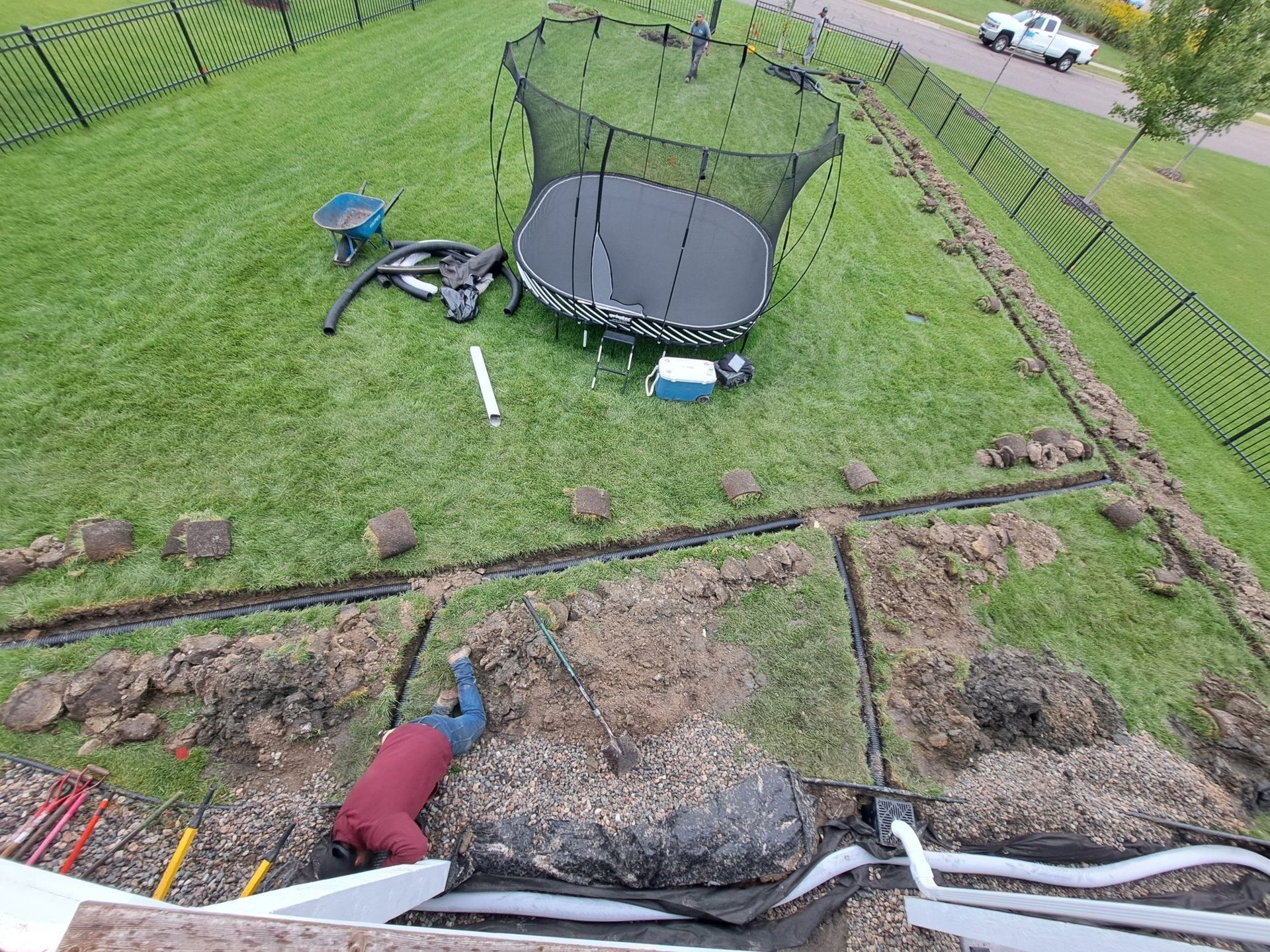 A high-angle view shows a person installing drainage pipes in a backyard, with trenches dug in the lawn near a trampoline.
