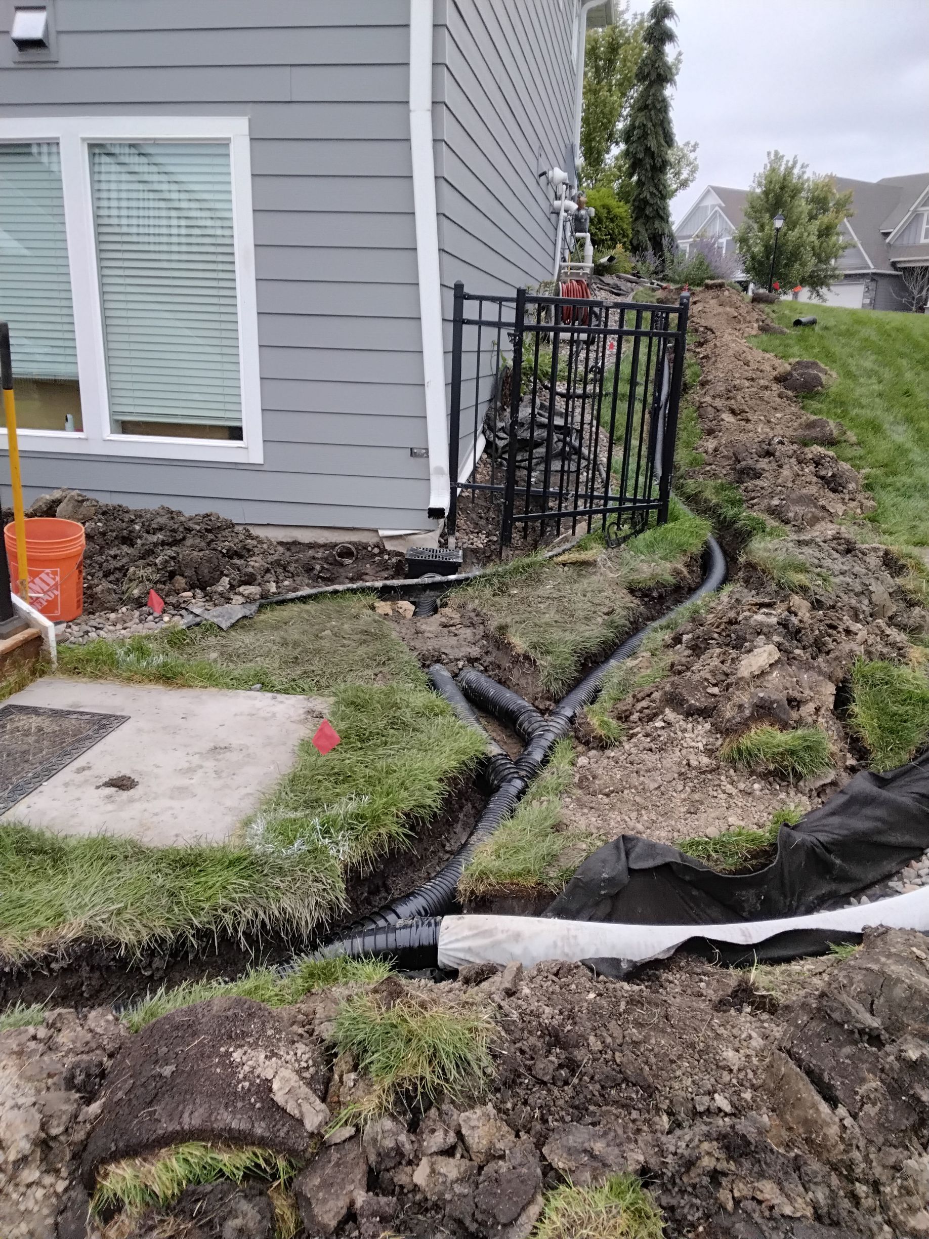 Black drainage pipes installed in a trench beside a grey house with a metal fence and landscaping debris.