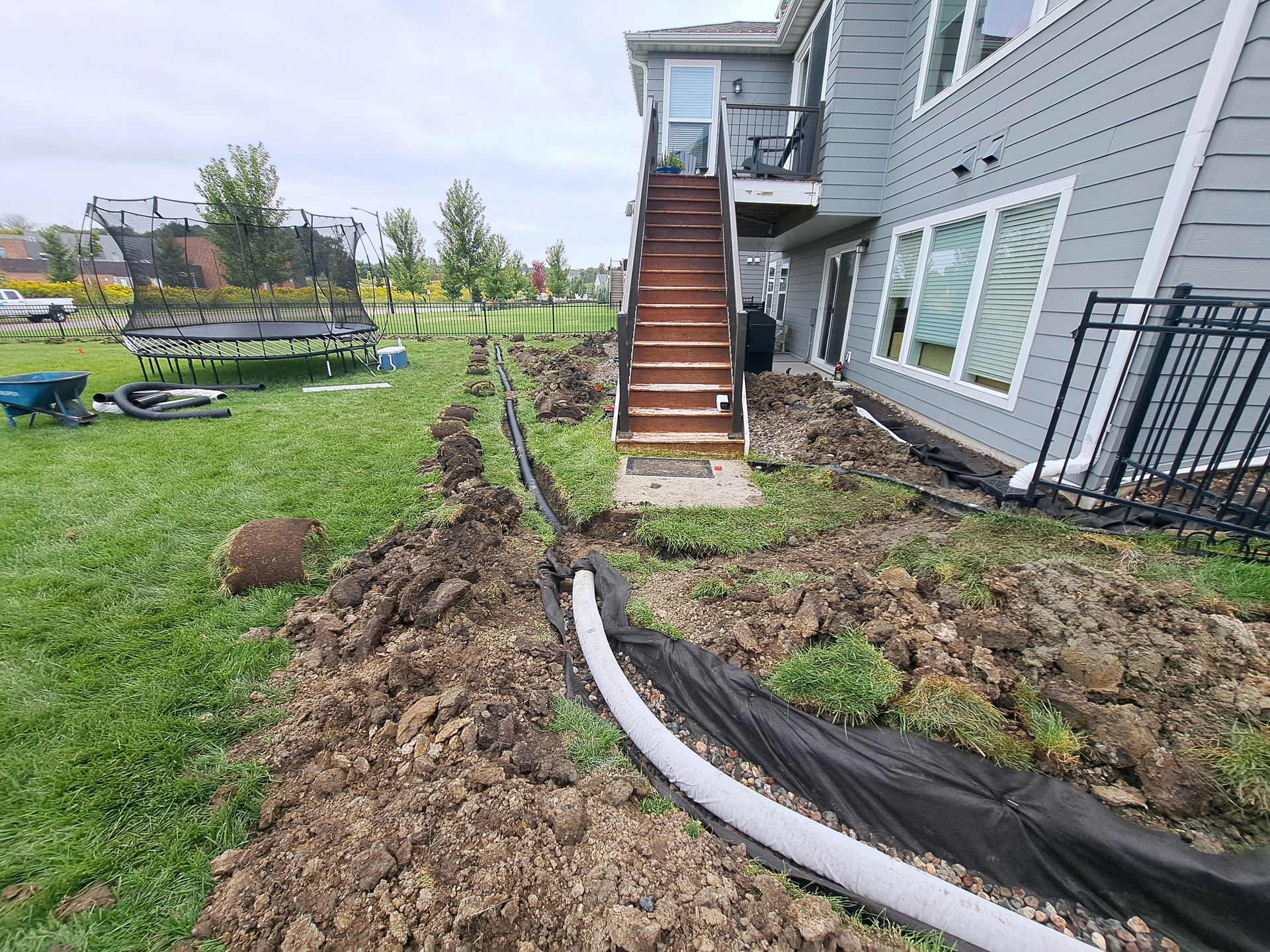 A trench with a white drainage pipe and landscaping fabric runs along the side of a house toward wooden stairs.