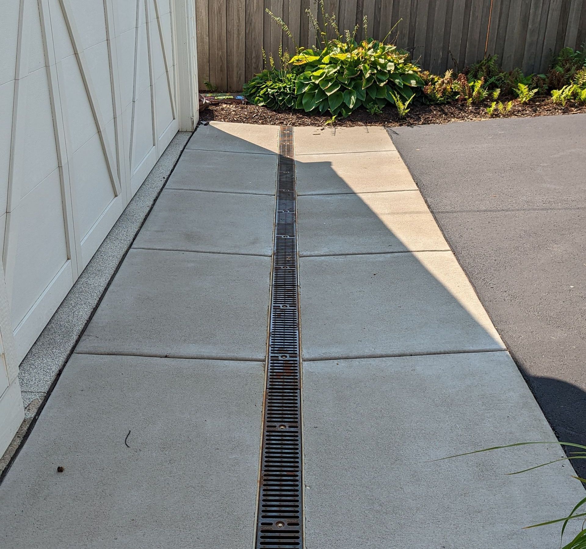 Concrete walkway with a central drain, next to a white fence and a black asphalt driveway.