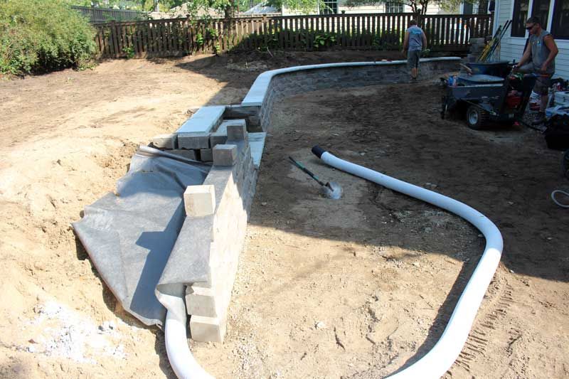 Construction of a retaining wall in a yard; workers, blocks, dirt, and drainage pipes are visible.