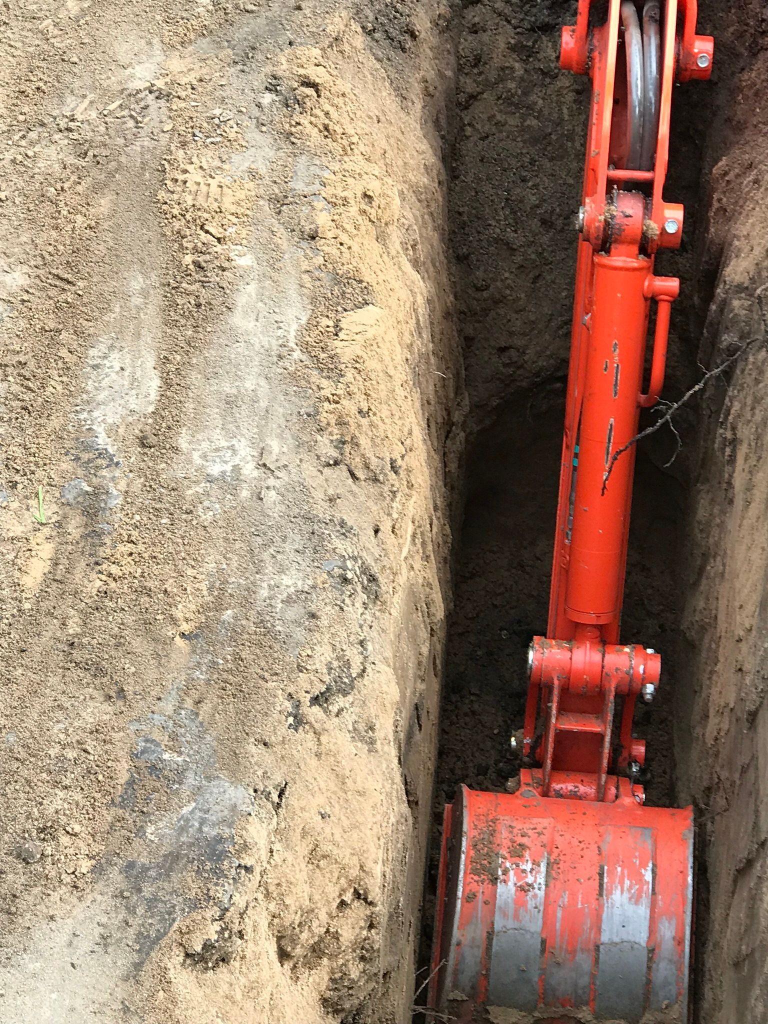Orange excavator bucket in a narrow trench, digging into brown soil.