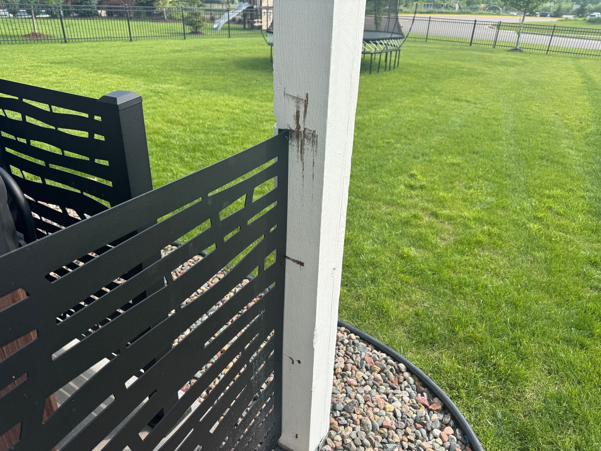 A decorative black metal privacy screen attached to a white wooden post outdoors with a green lawn and gravel bed nearby.