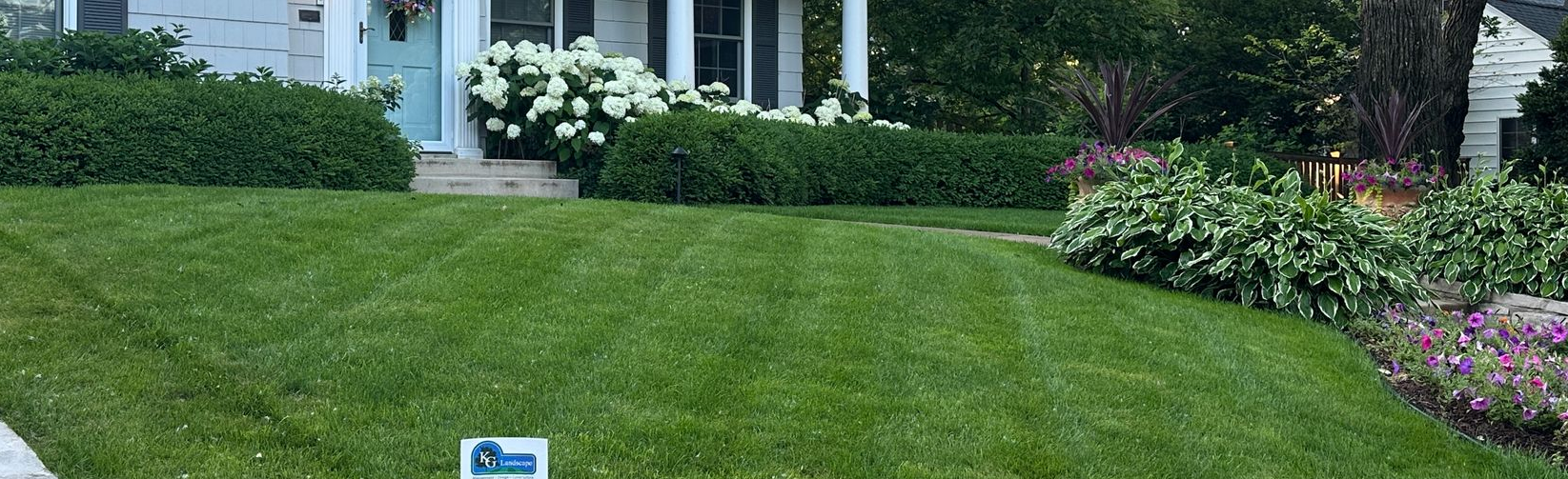 Lush green lawn with a brick pathway leading to a two-story gray house with a balcony and garden beds.