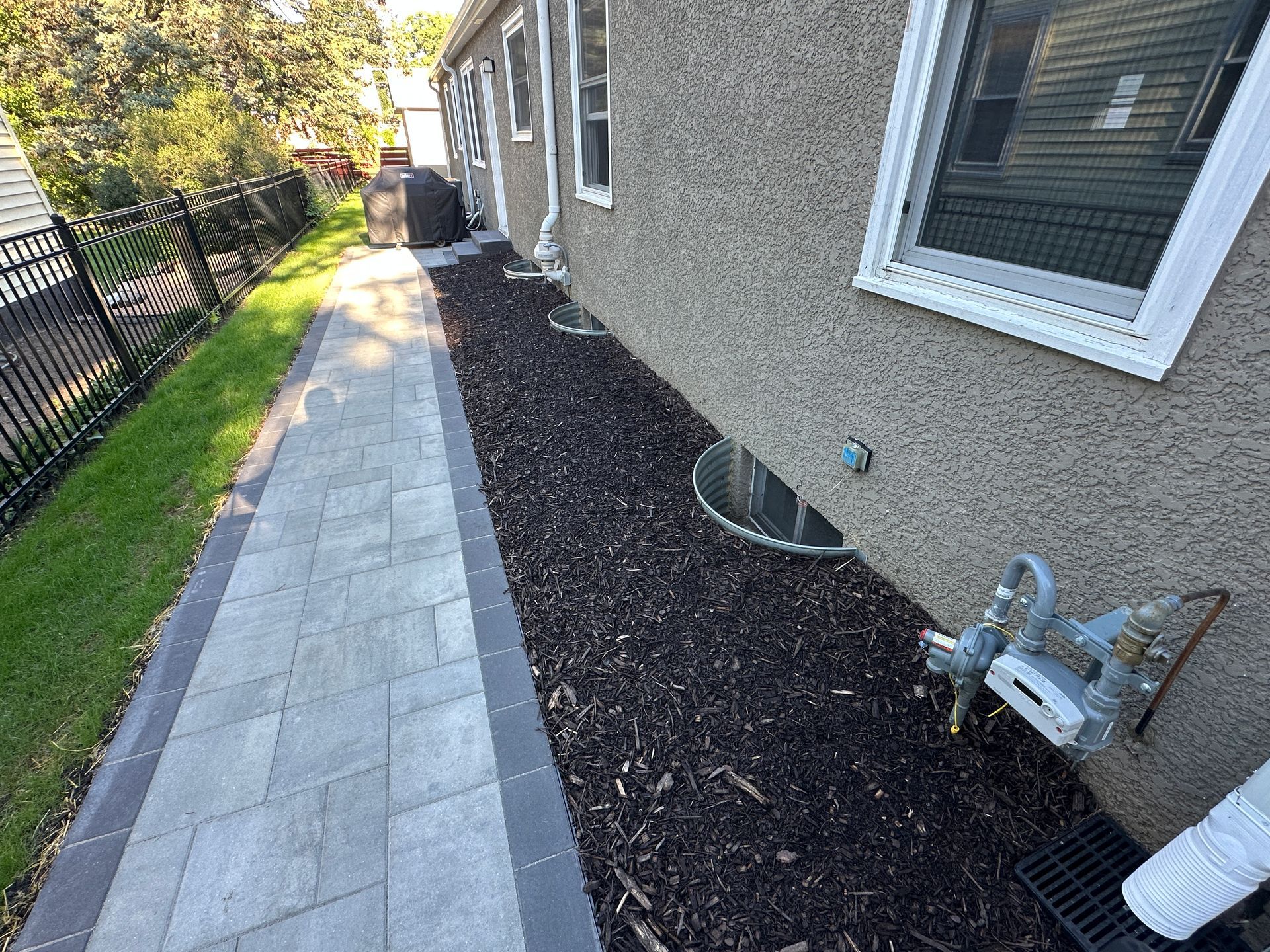 A paved stone walkway runs alongside a stucco house with dark mulch, basement window wells, and a gas meter.