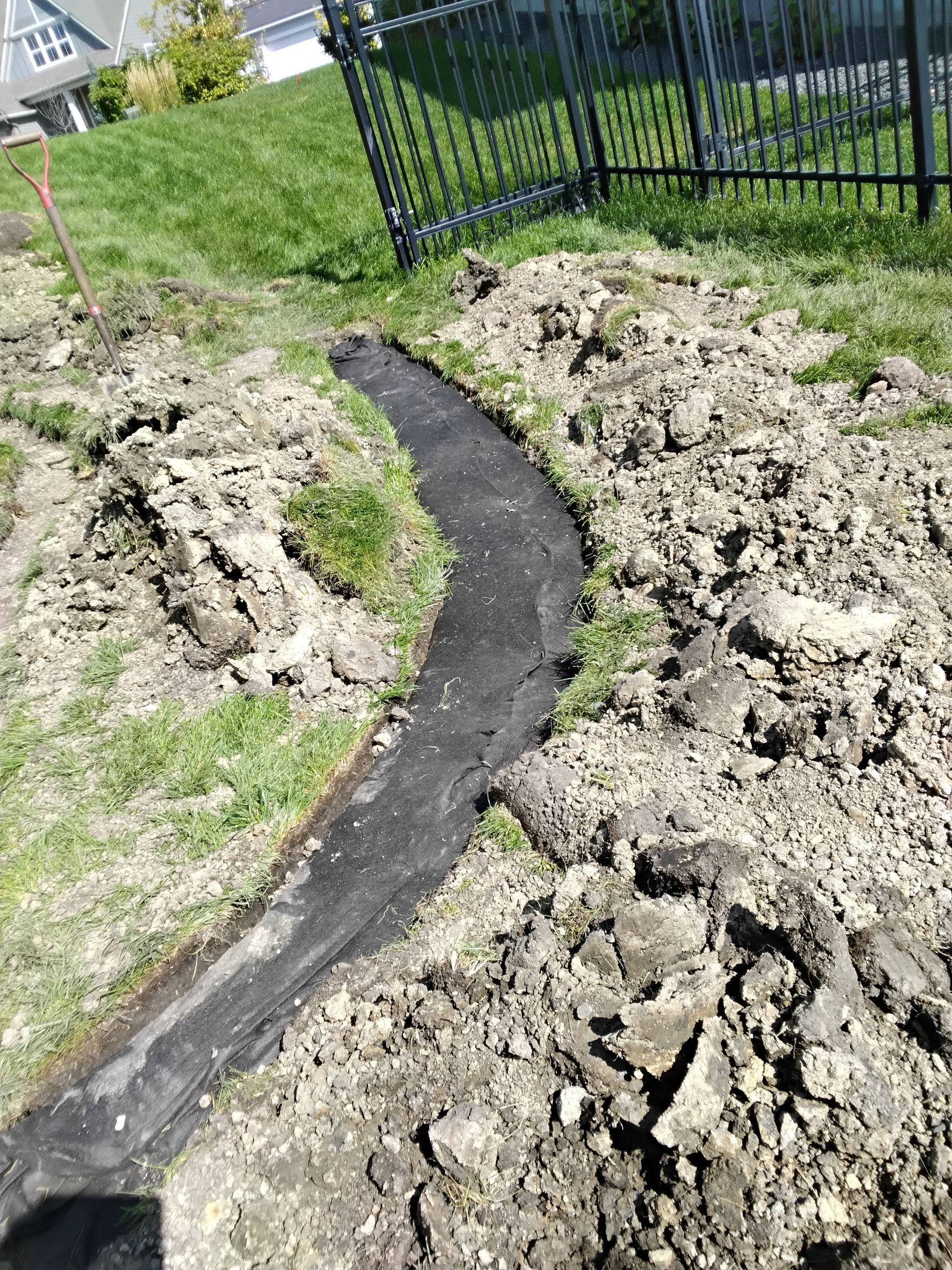 A black landscape fabric strip curves through a freshly dug dirt trench in a lawn near a black metal fence.