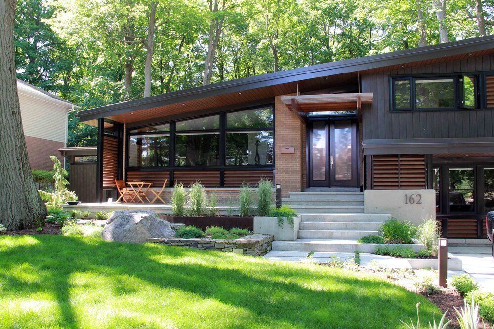 Modern, brown-sided house with angled roof, large windows, and manicured landscaping, front steps, and green lawn.