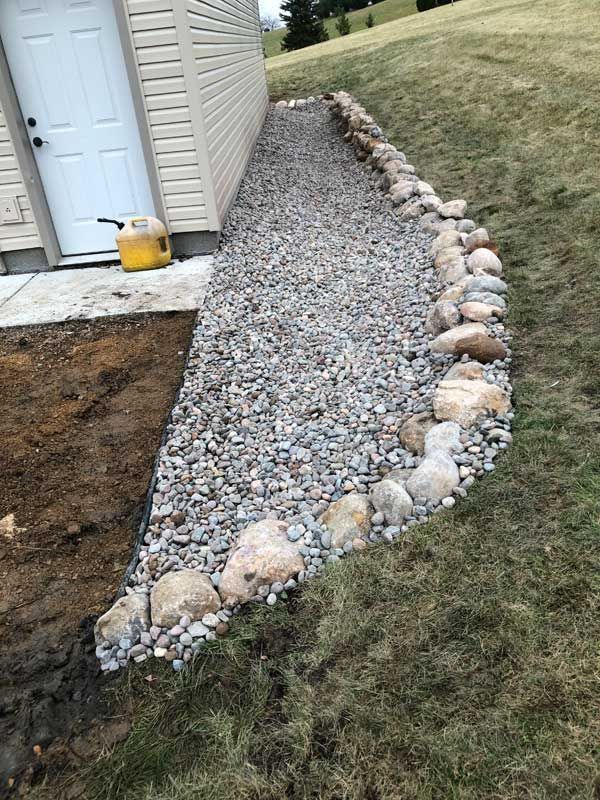 A gravel bed bordered by large rocks next to a building, with a grass slope on one side.