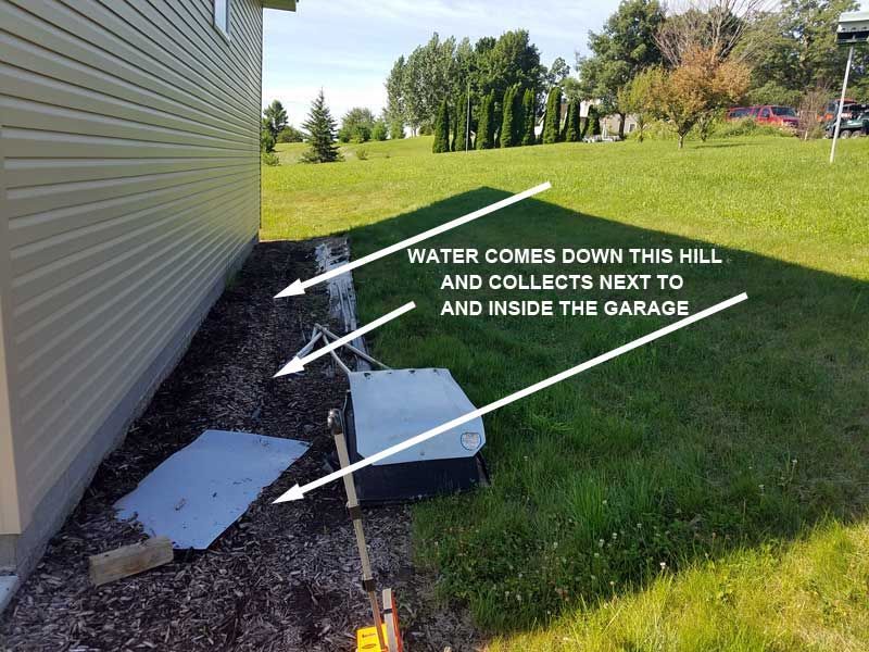 Water runoff collects near a garage. Gravel, grass, and a hill are present.