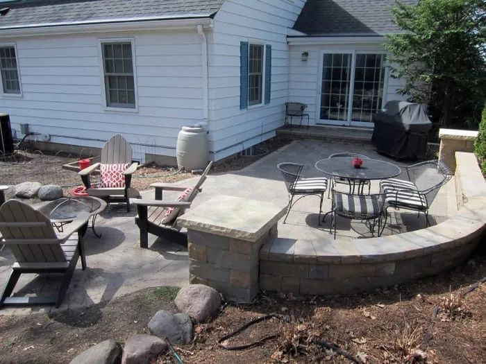 Backyard patio with fire pit, seating, and a low retaining wall by a white house with blue shutters.