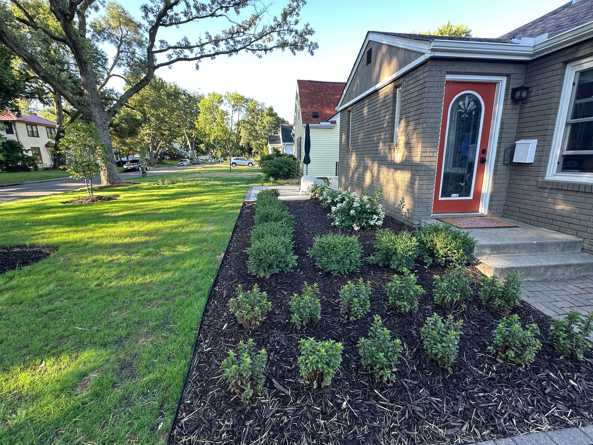 A house with dark brick siding and a bright orange front door, featuring a mulched front garden with small green shrubs.