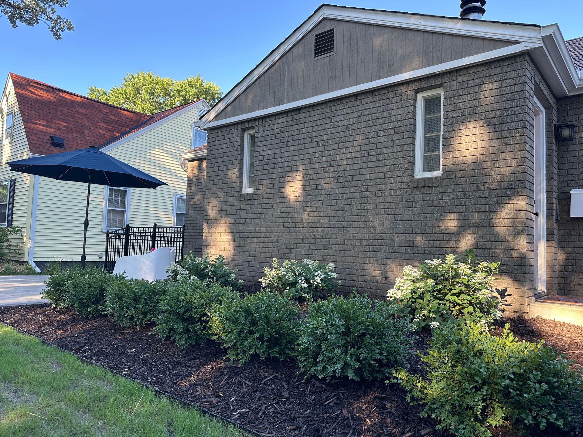 A brick house with a tan-sided home nearby, featuring a blue patio umbrella and green shrubs in a mulched garden bed.