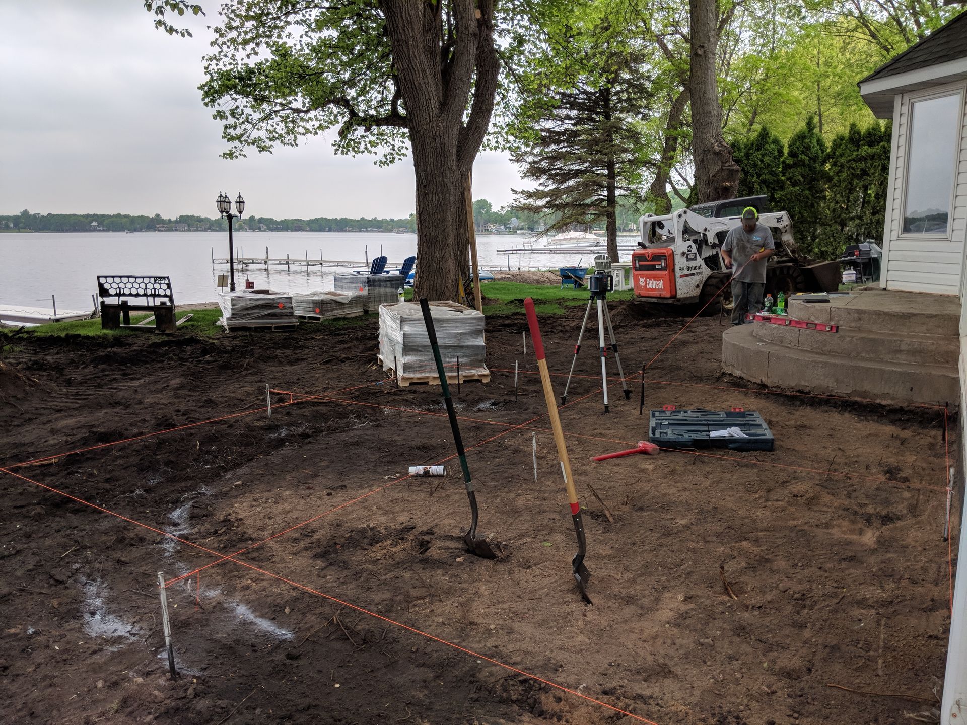 Construction site by a lake: Excavated ground with tools, a skid steer, and workers near a house.