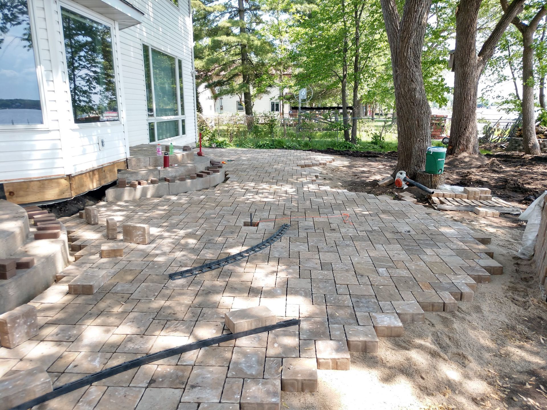 Brick pavers being laid on a patio near a white house and trees.