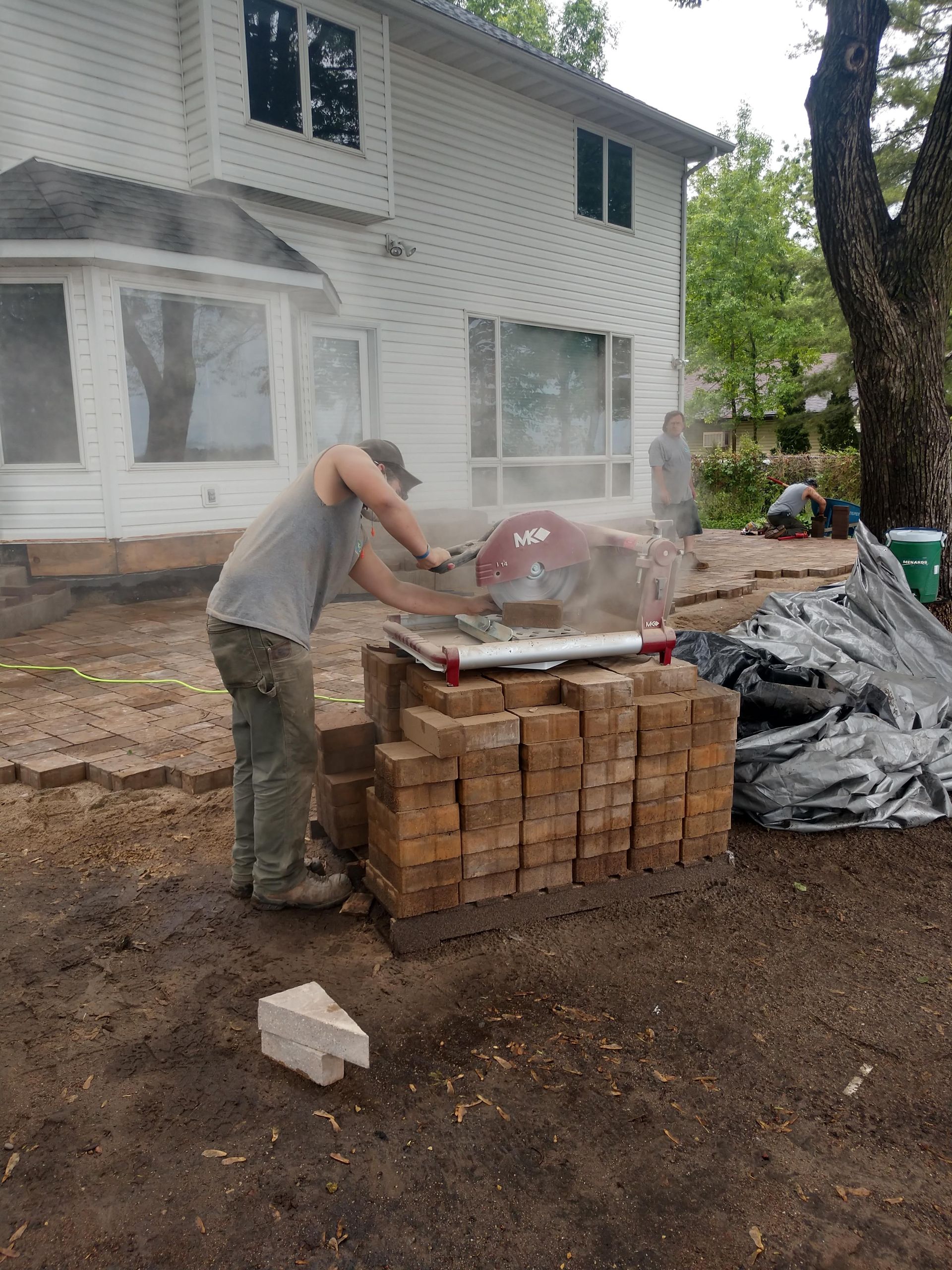 Man using a saw to cut bricks outside a white house. Dust fills the air.