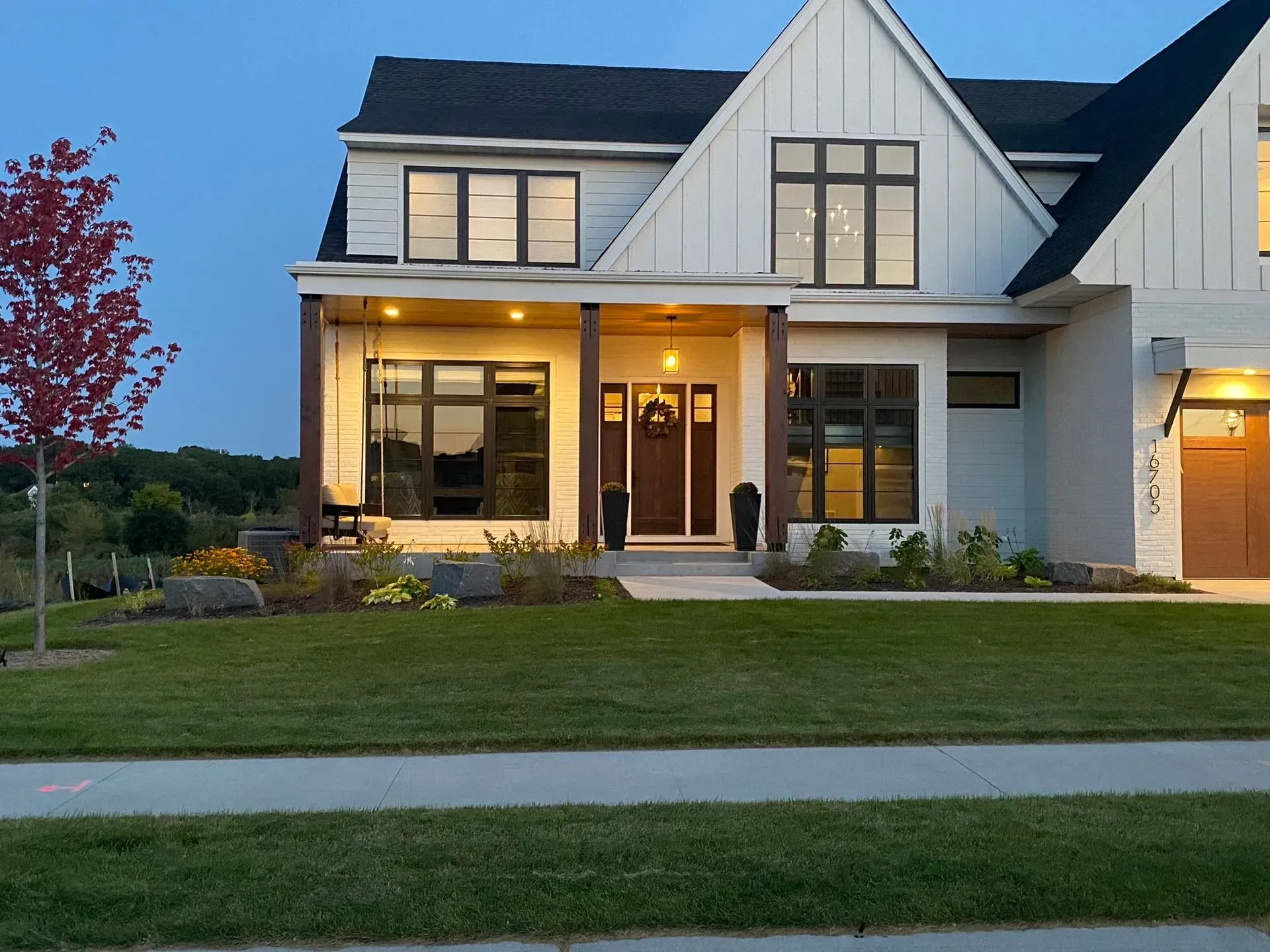 White two-story house with dark windows, brown front door, lit porch, and manicured lawn.