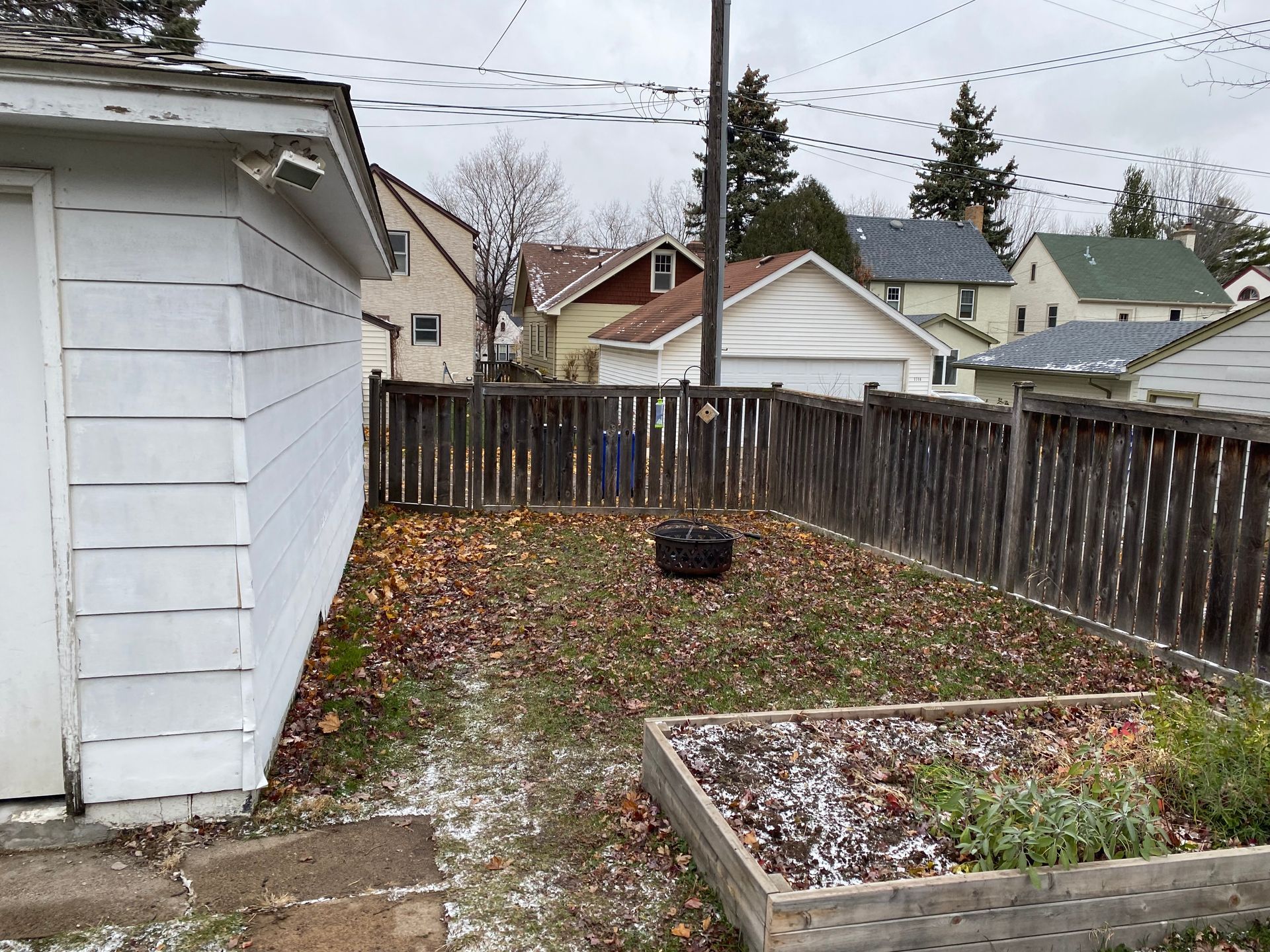 A fenced-in backyard with a white garage, scattered autumn leaves, light snow, and a raised wooden garden bed.