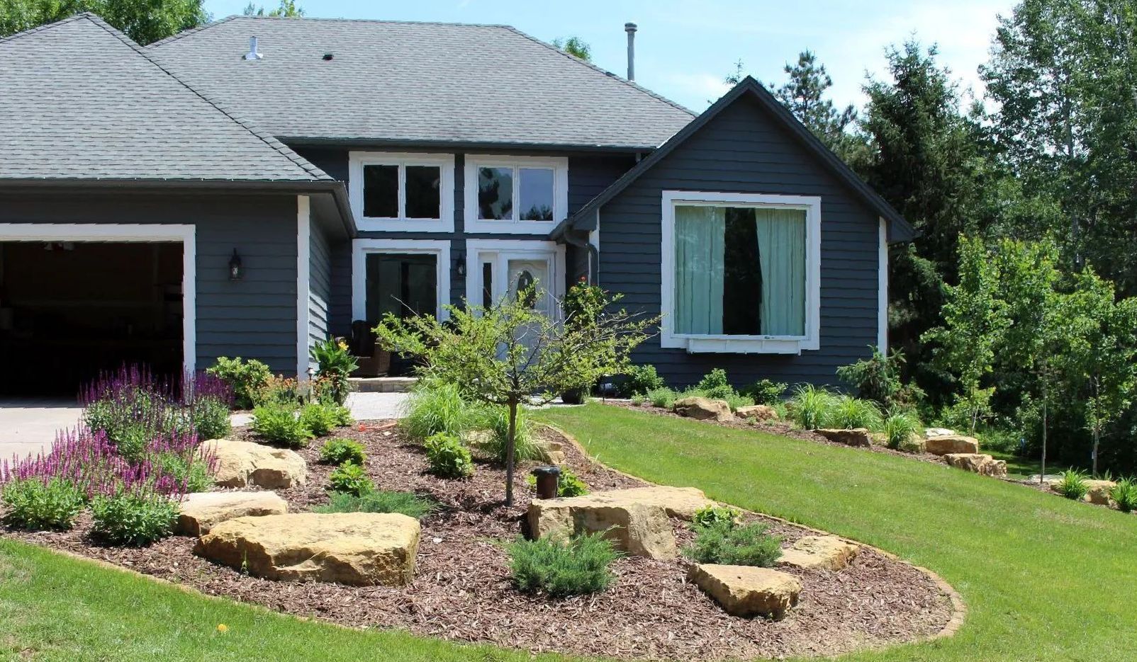 Sloped front yard with boulder outcroppings, Tina crab focal tree, curved stone-edged bed, and brown mulch, Ham Lake MN