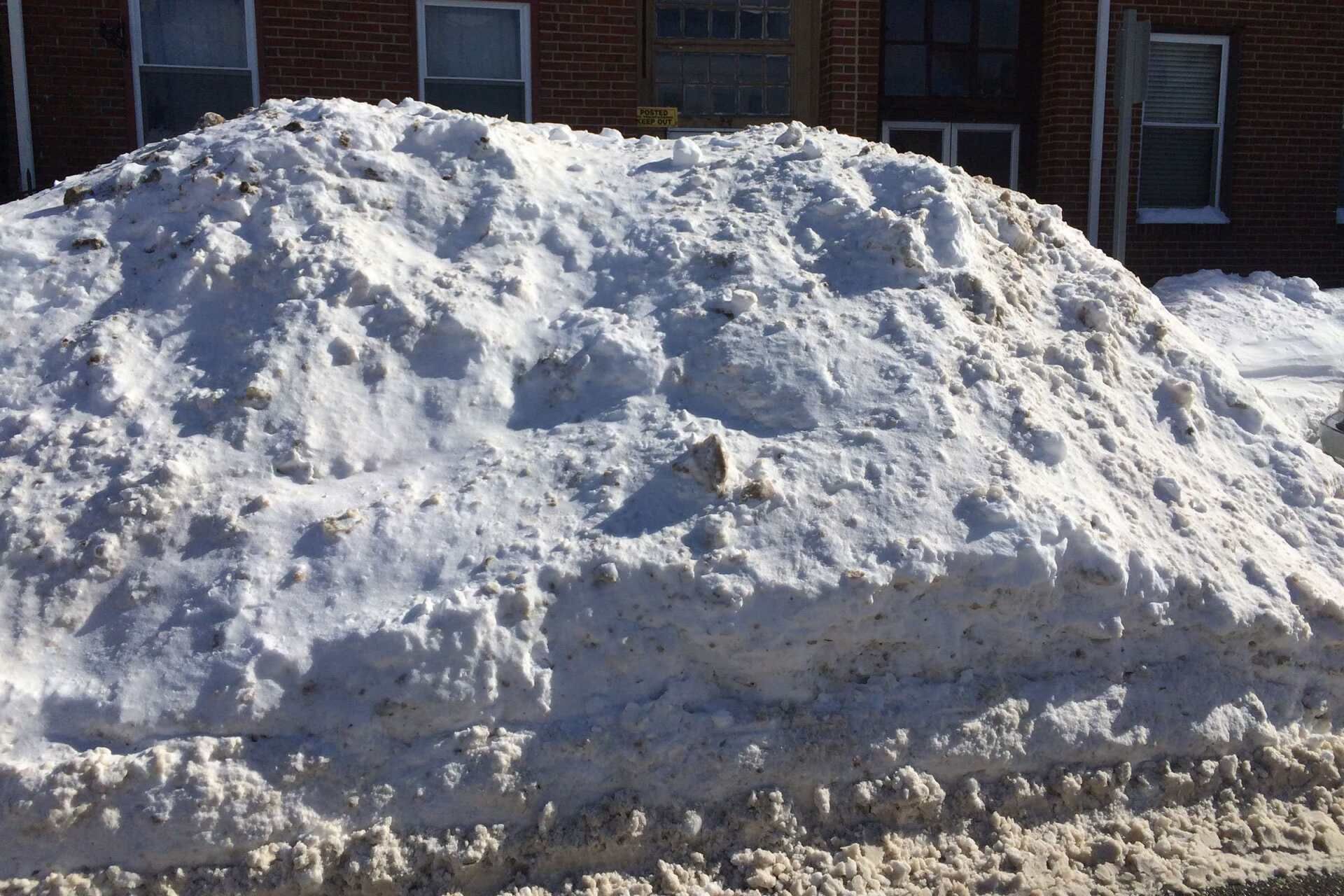 A large, dirty snowbank piled up in front of a brick building.