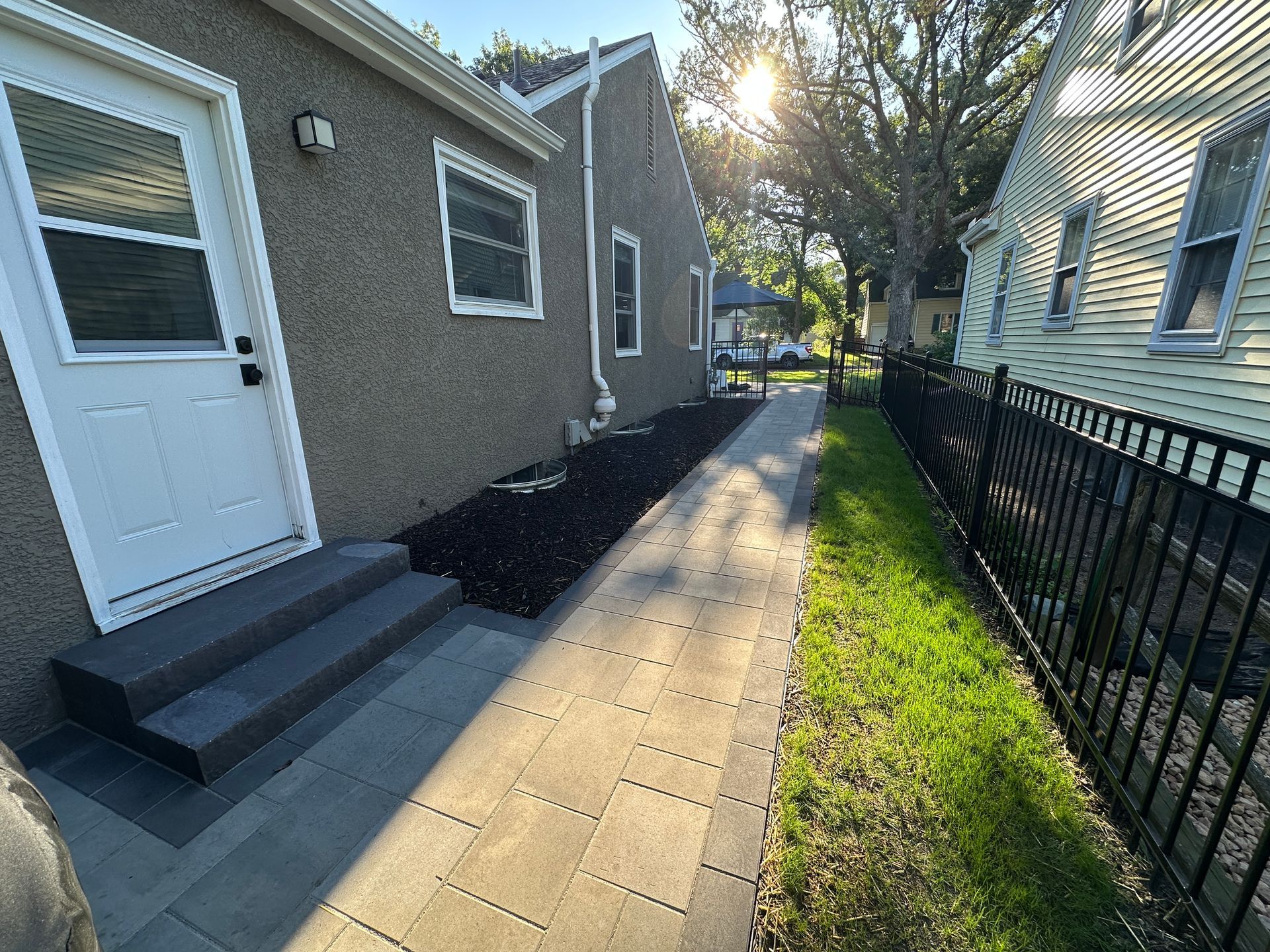 A gray stucco house with a white door and stone steps leading to a paved walkway beside a black metal fence and lawn.