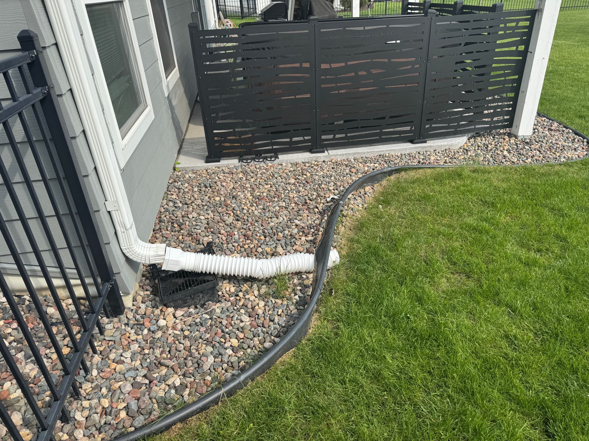A white downspout extension leads into a black catch basin near a gray house with a black privacy screen and gravel border.