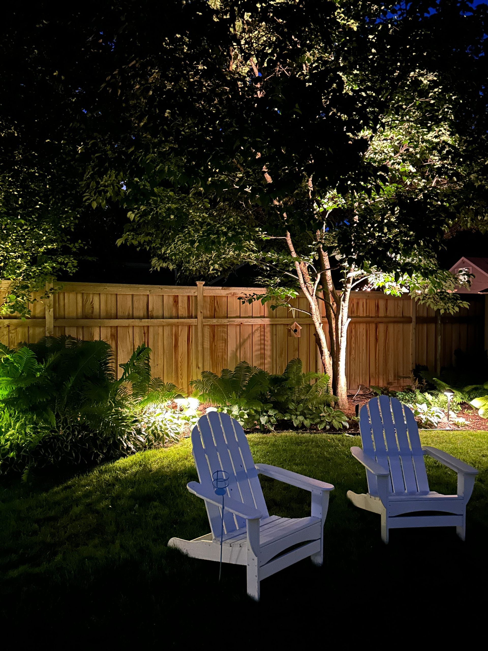 Two white Adirondack chairs on a lit lawn at night, beneath a tree and fence.