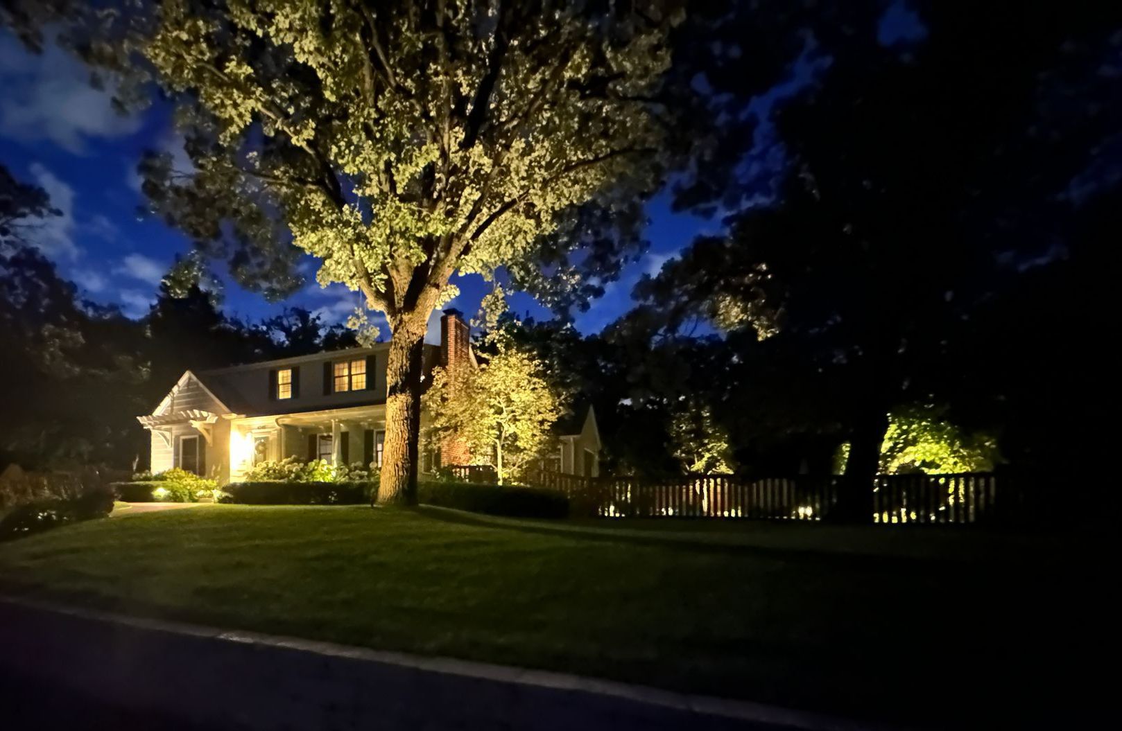 House lit up at night with landscape lighting, trees, and lawn under a twilight sky.