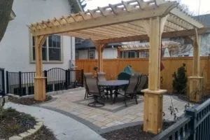 Wooden pergola over a patio with a table and chairs, near a house and fence.