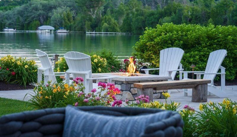 Lakeside fire pit with white Adirondack chairs, bench, and colorful flowers; lake in background.